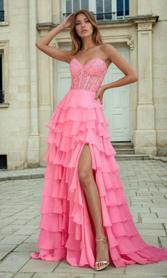 A woman in the PromGirl 250001 Strapless Long Ruffled Prom Gown with sheer corset bodice and high slit stands outdoors on a stone walkway before a beige building with tall doors.