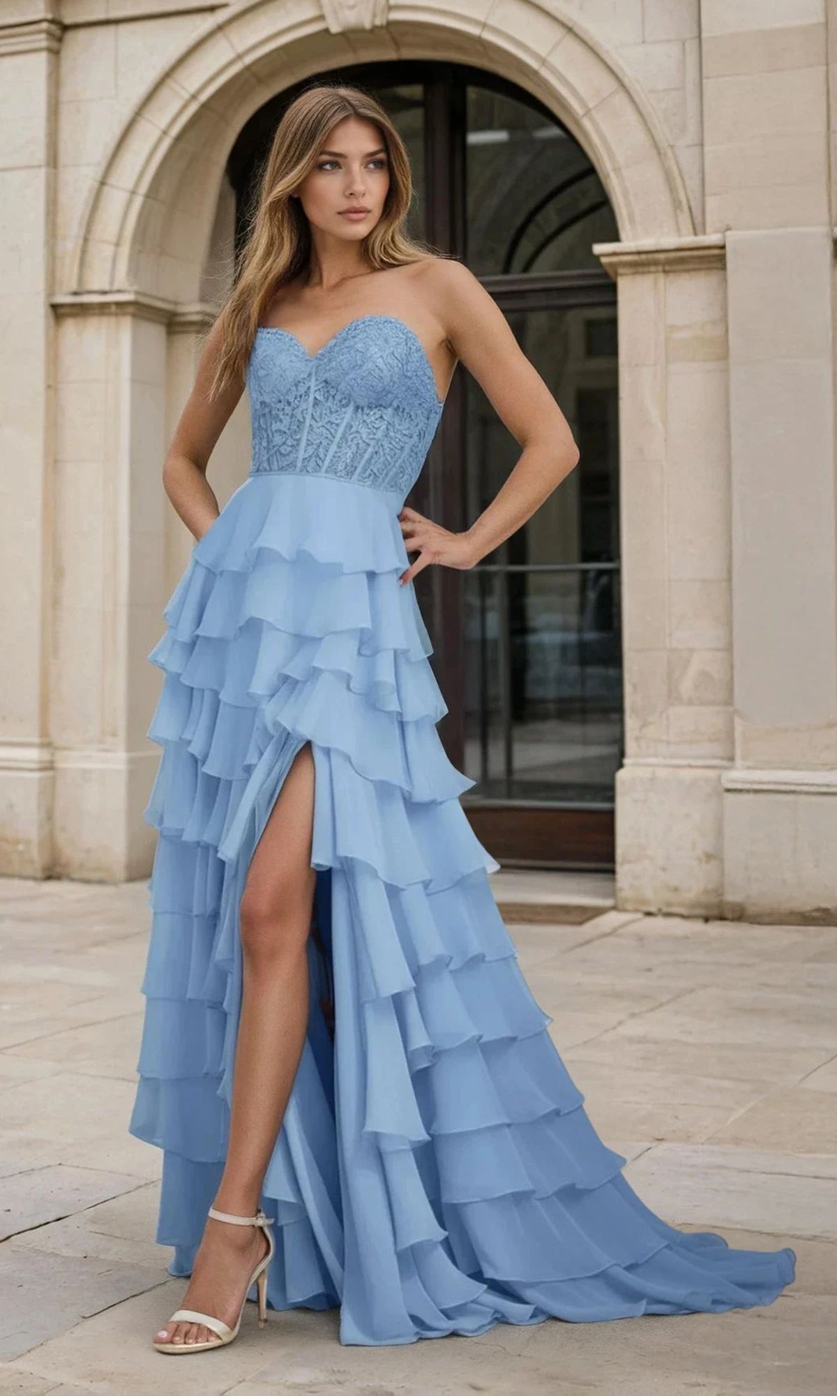 A woman stands outdoors in front of an arched doorway, wearing the PromGirl 250001 Strapless Long Ruffled Prom Gown with a sheer corset bodice, light blue lace details, tiered skirt, and front slit.