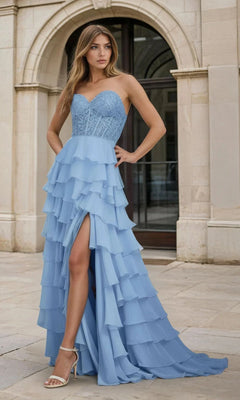 A woman stands outdoors in front of an arched doorway, wearing the PromGirl 250001 Strapless Long Ruffled Prom Gown with a sheer corset bodice, light blue lace details, tiered skirt, and front slit.