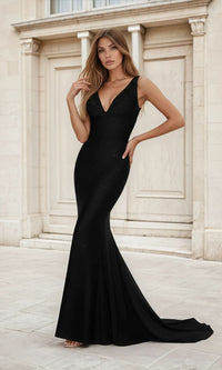 A woman stands outdoors before a stone building, wearing the PromGirl Exclusive Empire-Waist Mermaid Prom Dress—a fitted, floor-length black gown with a deep V-neckline.