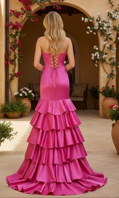 Wearing the PromGirl Exclusive 250069 long prom dress, a woman stands outdoors in a strapless mermaid gown with a lace-up back, her bright pink ruffled skirt glowing among potted plants and blooming vines.