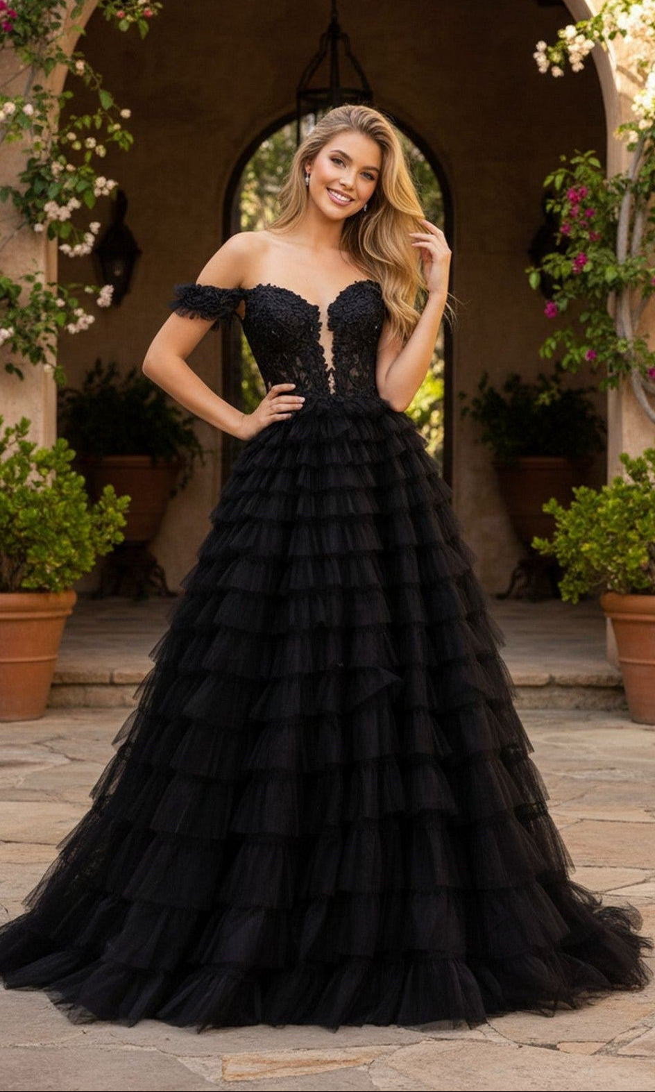 A woman smiles and poses outdoors in the PromGirl Exclusive 250070, a black strapless prom ball gown featuring a lace corset bodice and tiered ruffle tulle skirt, with one hand near her hair.