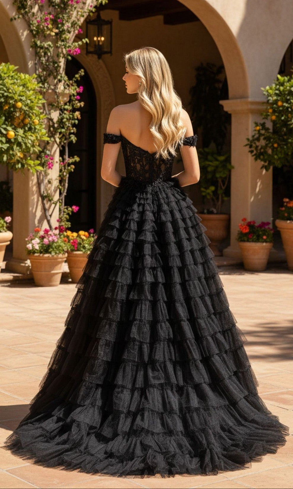 A woman in the PromGirl Exclusive 250070 black off-the-shoulder ball gown with a lace corset bodice stands outdoors on a tiled patio, surrounded by potted plants and arched architecture.