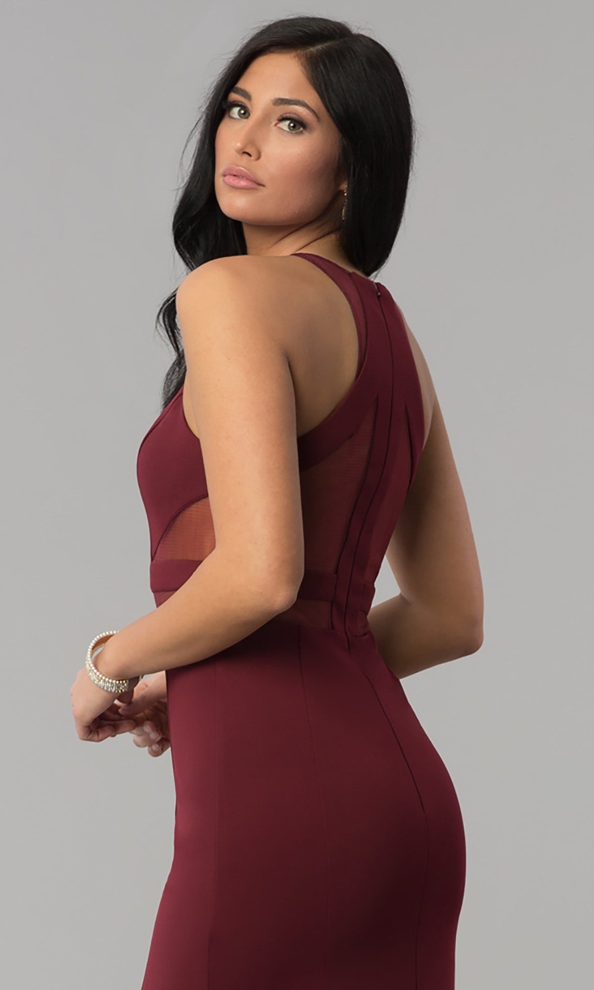 A woman with long dark hair wears the Illusion-Cut-Out High-Neck Long Prom Dress 8054, a sleeveless maroon gown with sheer side panels, against a plain gray background.