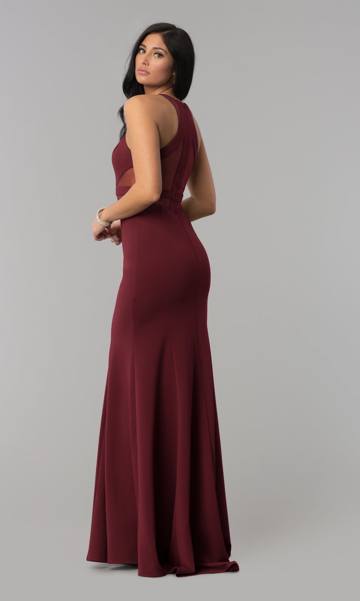 A woman stands against a plain gray background, looking over her shoulder while wearing the Illusion-Cut-Out High-Neck Long Prom Dress 8054—a sleeveless, floor-length burgundy bodycon gown.
