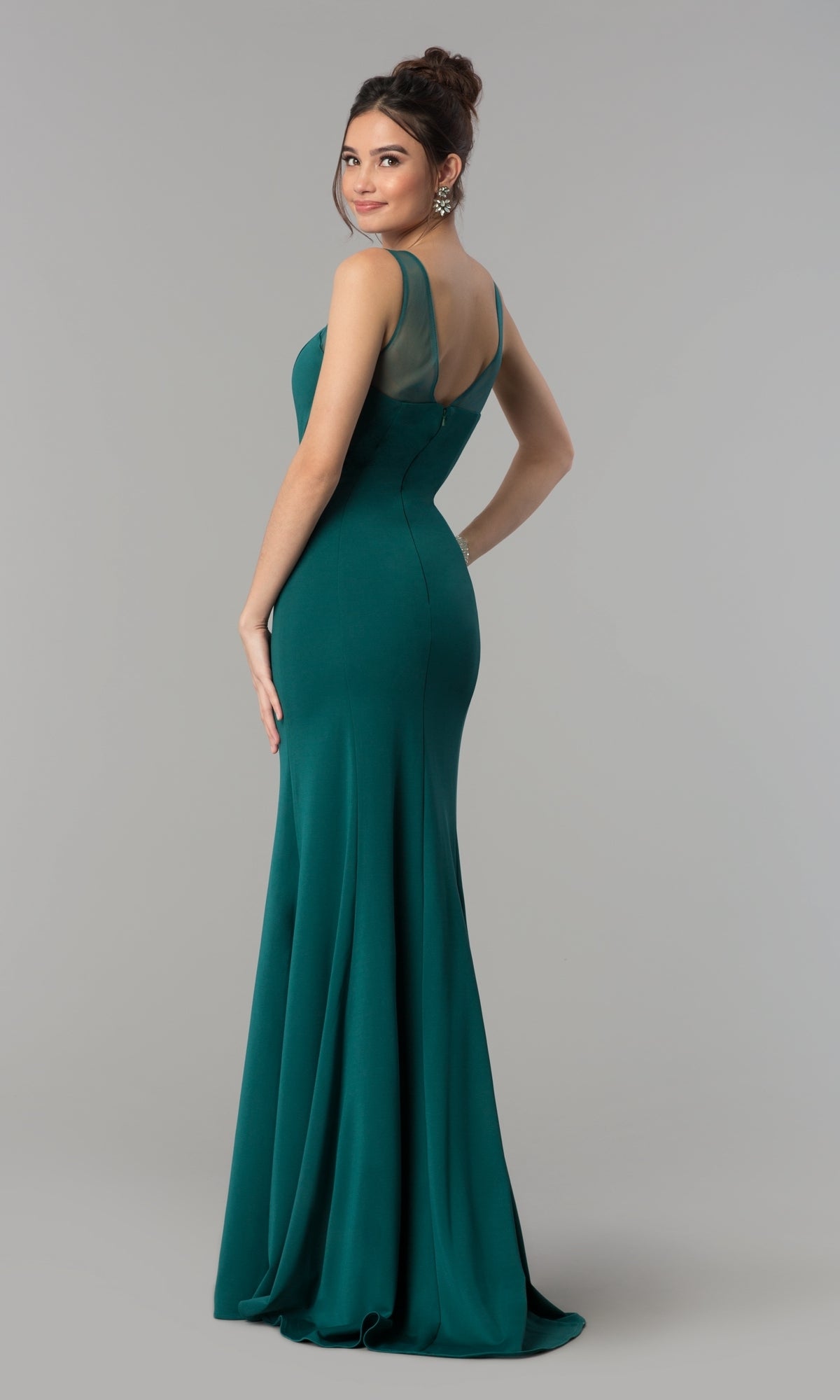 Woman in the Illusion-Neckline Long Jersey Formal Prom Dress 8148, a sleeveless, floor-length teal gown with sheer shoulder panels, stands and looks over her shoulder against a plain gray background.