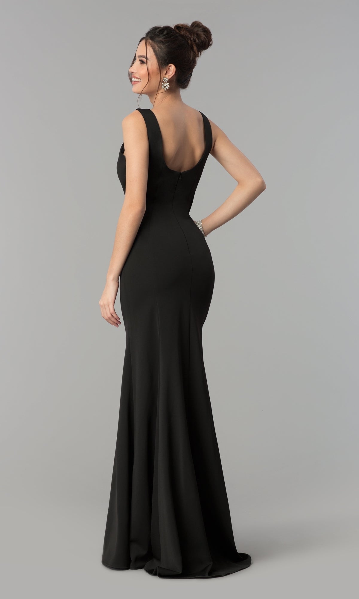 A woman in the Simple V-Neck Long Formal Jersey Prom Dress 8152, a floor-length, sleeveless black bodycon gown with a deep V-back, stands looking over her shoulder against a plain gray background.