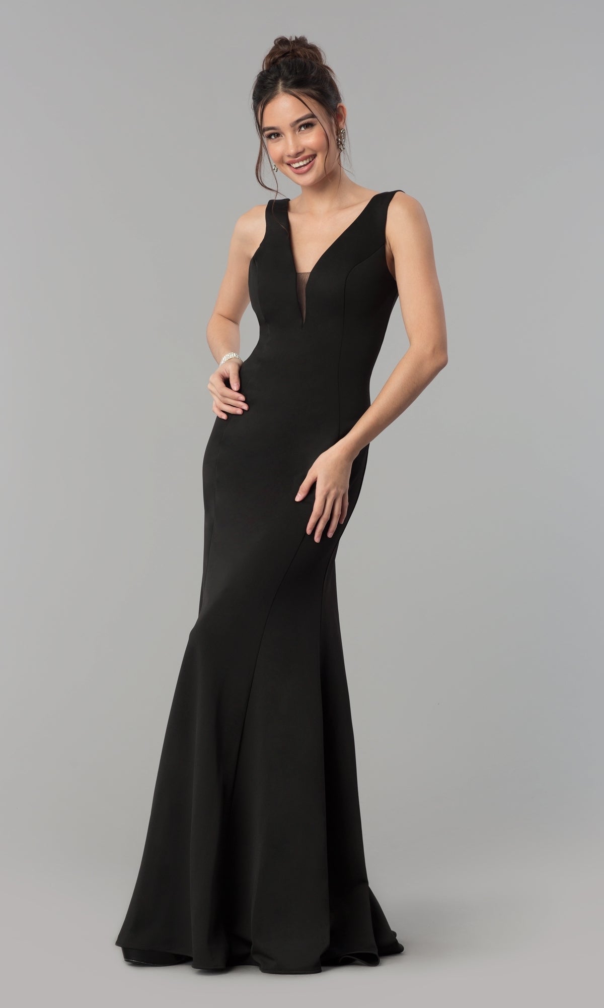 A woman stands and smiles in the Simple V-Neck Long Formal Jersey Prom Dress 8152 in black, featuring a sleeveless, floor-length design against a plain gray background.