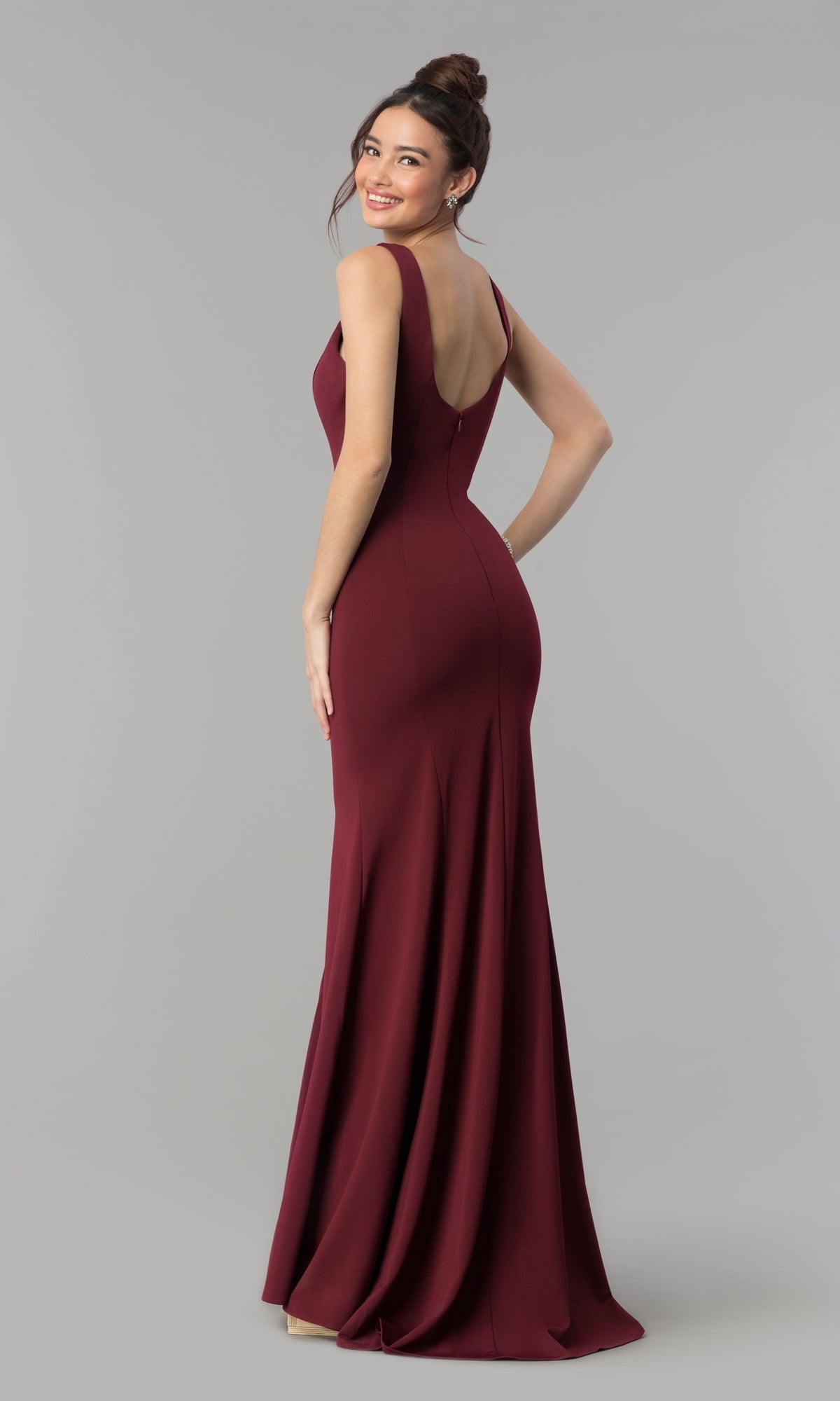 Woman models the Simple V-Neck Long Formal Jersey Prom Dress 8152 in burgundy, featuring a sleeveless v-neck design, as she smiles over her shoulder against a plain grey background.