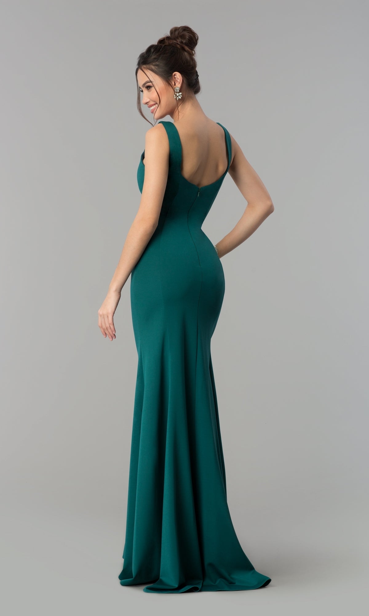 A woman in the Simple V-Neck Long Formal Jersey Prom Dress 8152, a sleeveless, floor-length emerald green bodycon gown, stands facing away and looks over her shoulder against a plain gray background.