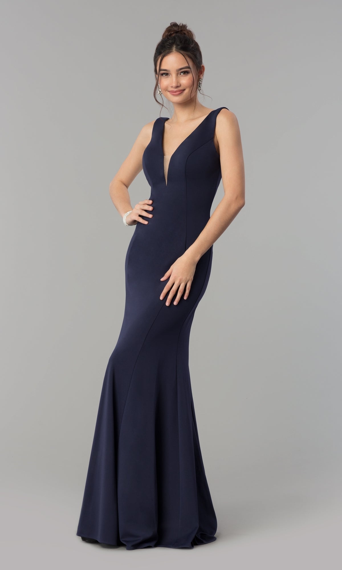 A woman poses against a plain gray background wearing the Simple V-Neck Long Formal Jersey Prom Dress 8152, a sleeveless, floor-length navy blue gown with a deep V neckline.