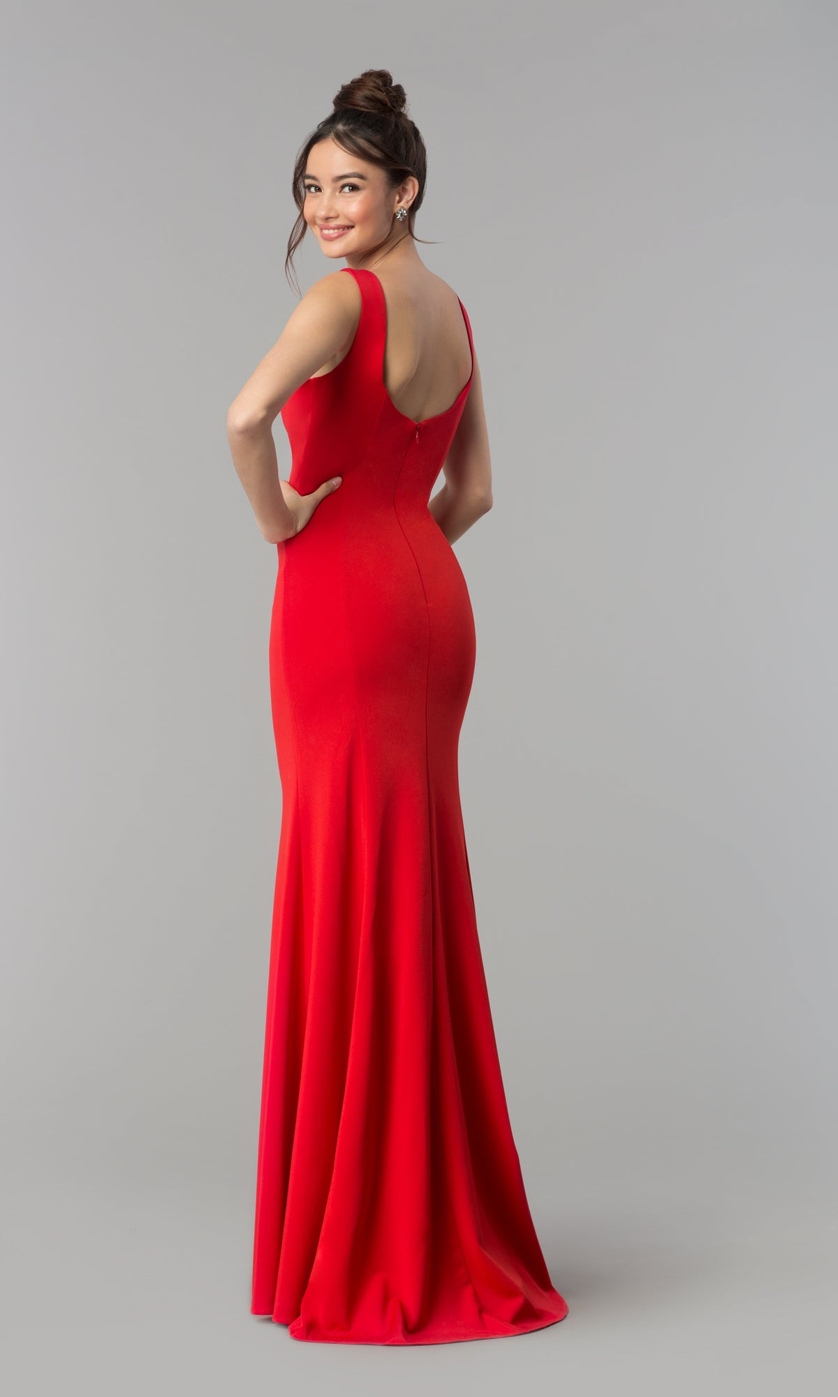 A woman faces backward, smiling over her shoulder in the Simple V-Neck Long Formal Jersey Prom Dress 8152, a floor-length, sleeveless red gown, set against a plain light gray background.