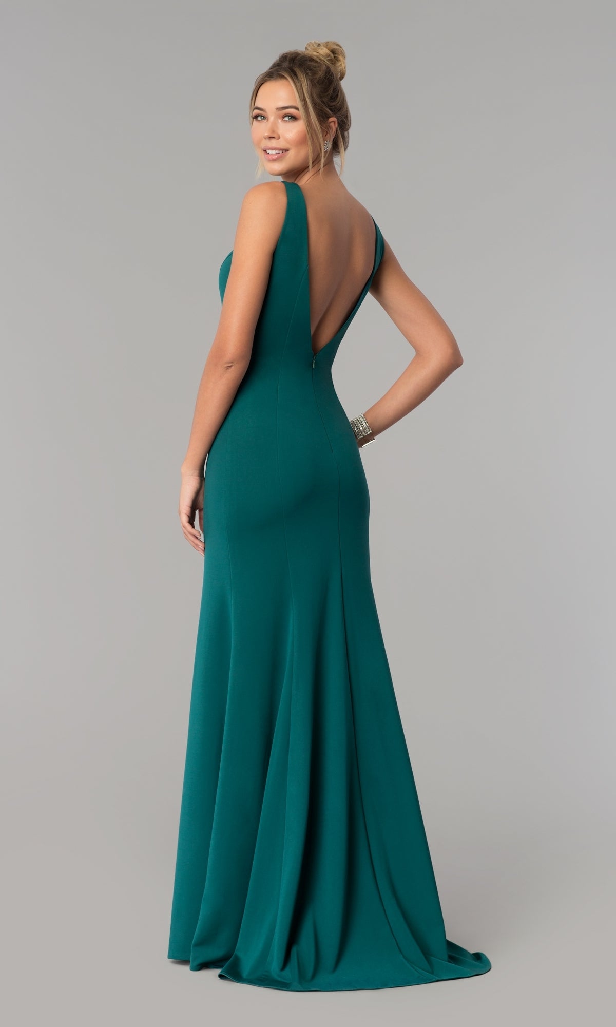 A woman stands against a plain gray background, looking over her shoulder while wearing the Sleeveless V-Neck Long Formal Prom Dress 8158 in emerald green, featuring a floor-length skirt and a deep V-back.