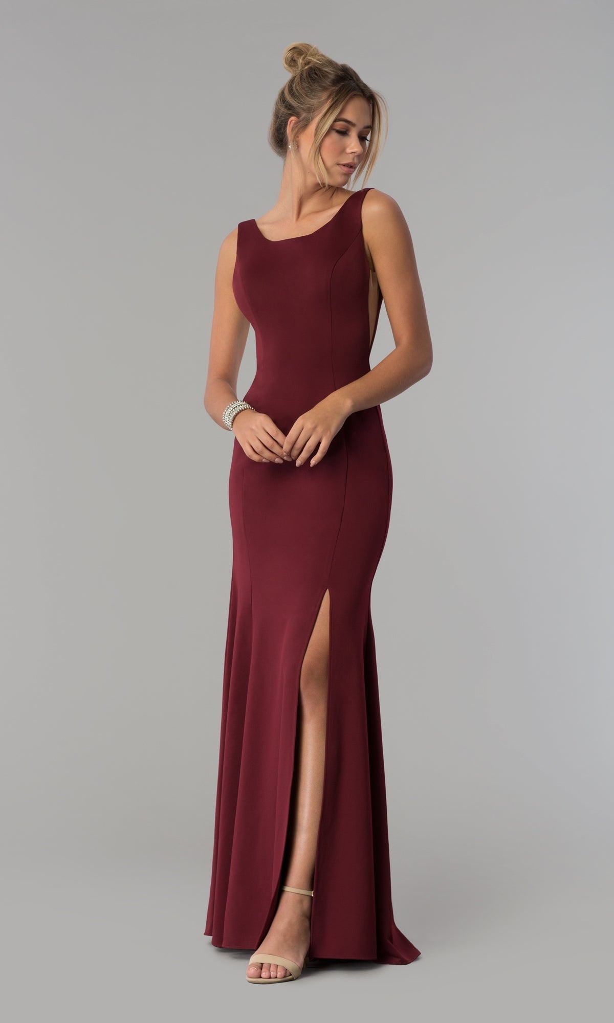 A woman wears the Sheer-Sided Backless Long Formal Prom Dress 8168 in burgundy with a sleeveless, floor-length design and thigh-high slit, paired with nude heels, standing against a plain gray background.