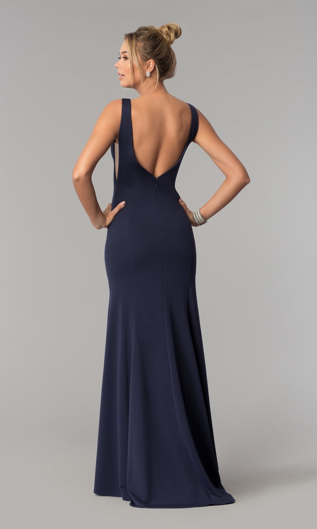 A woman stands with hands on hips, facing away, in the Sheer-Sided Backless Long Formal Prom Dress 8168— a sleeveless navy floor-length gown with a deep V-back—against a plain gray background.