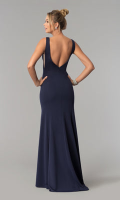 A woman stands with hands on hips, facing away, in the Sheer-Sided Backless Long Formal Prom Dress 8168— a sleeveless navy floor-length gown with a deep V-back—against a plain gray background.