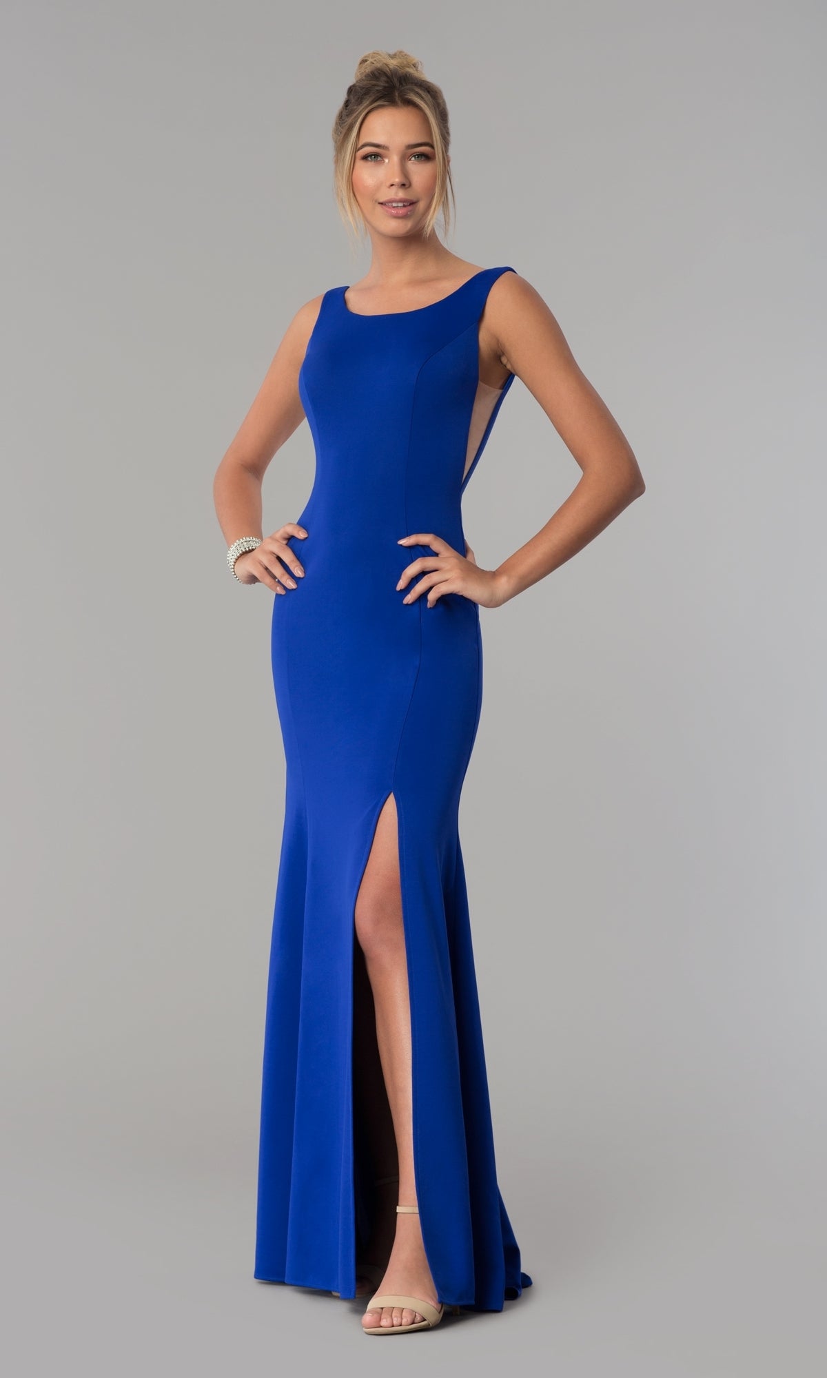 A woman poses against a gray background wearing the Sheer-Sided Backless Long Formal Prom Dress 8168, a sleeveless, floor-length royal blue jersey gown with a high slit.