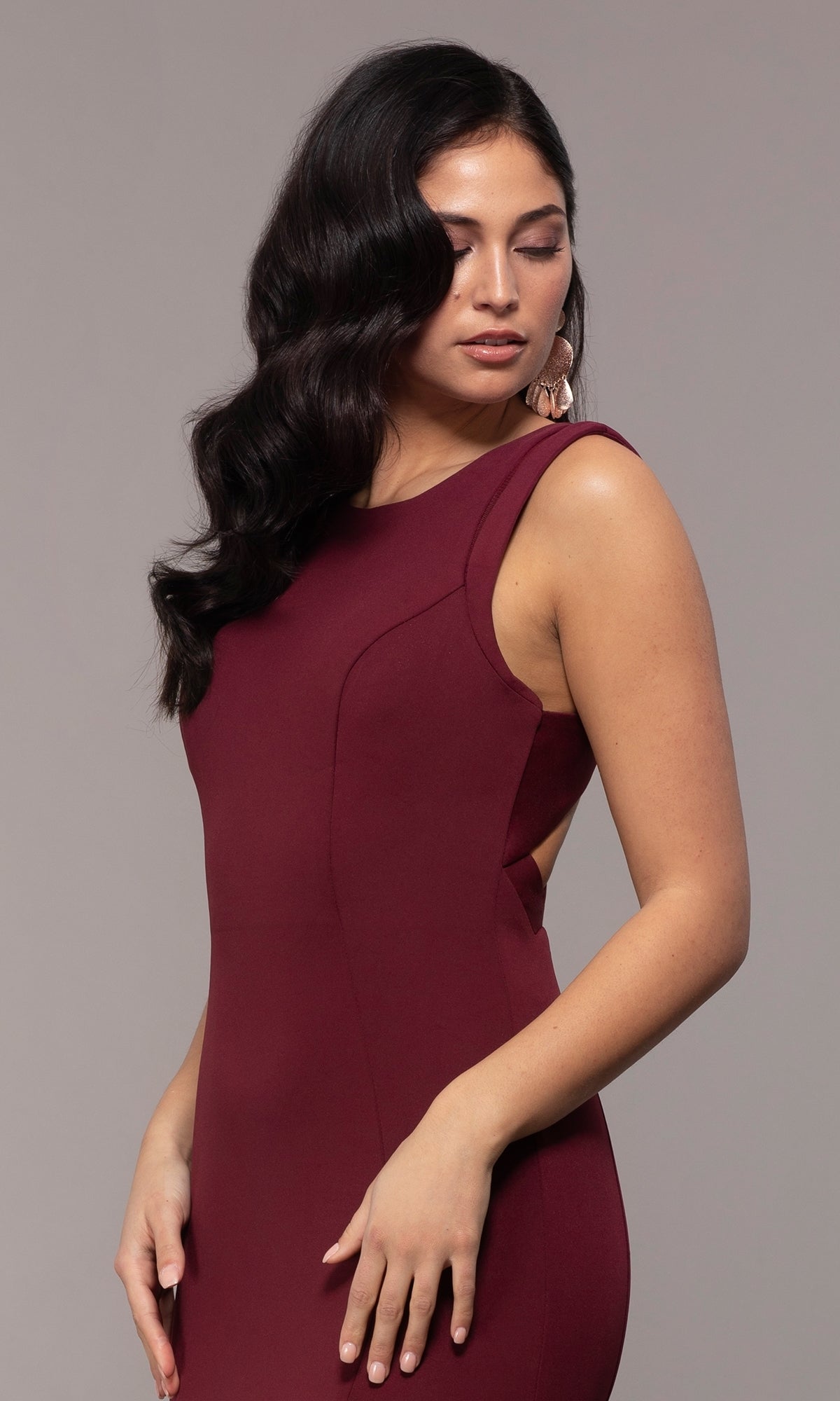 Woman with long dark wavy hair models the Strappy-Open-Back Long Formal Prom Dress 8232, posing against a plain gray background, looking down with one hand resting at her side.