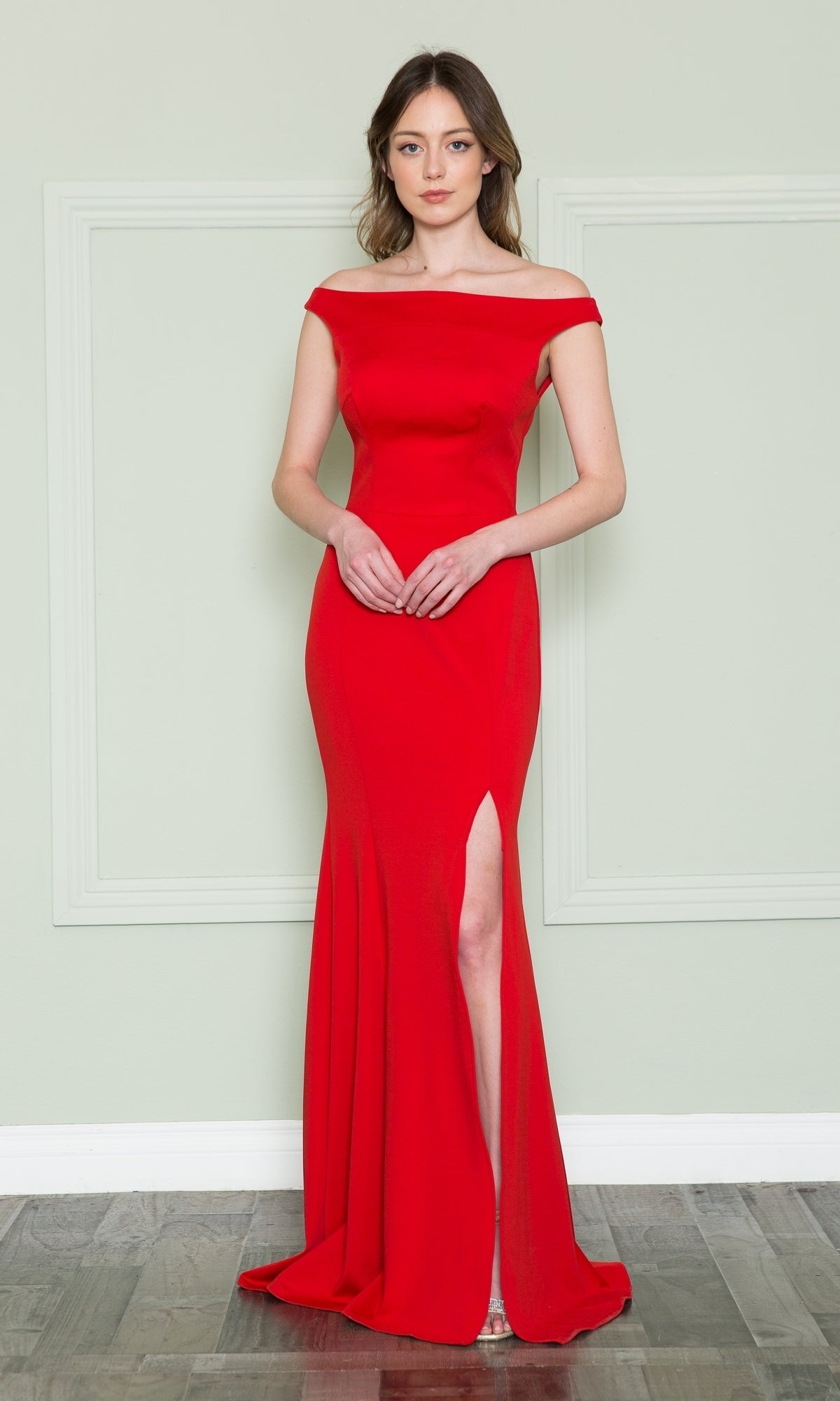 A woman stands against a light-colored wall wearing the Off-the-Shoulder Classic Long Prom Dress 8724 in red, featuring a fitted silhouette and thigh-high slit, with her hands clasped in front.