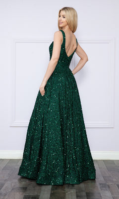 A woman in the Velvet-Sequin Long A-Line Prom Gown (Poly USA 9106), a sleeveless, floor-length dark green dress, stands with her back turned and looks over her shoulder in a minimalist indoor setting.