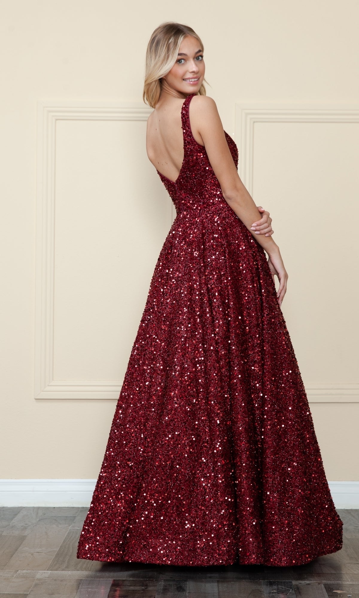 Woman in a Velvet-Sequin Long A-Line Prom Gown (Poly USA 9106) poses with her back slightly turned, standing indoors against a beige wall with decorative molding.
