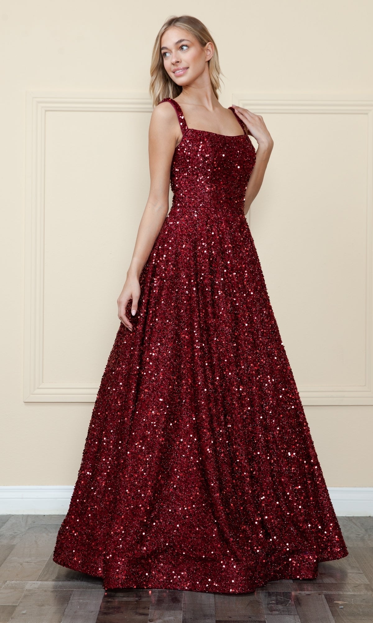 A woman stands indoors against a beige wall, wearing the Poly USA 9106 Velvet-Sequin Long A-Line Prom Gown—a floor-length, sleeveless red dress with a square neckline.