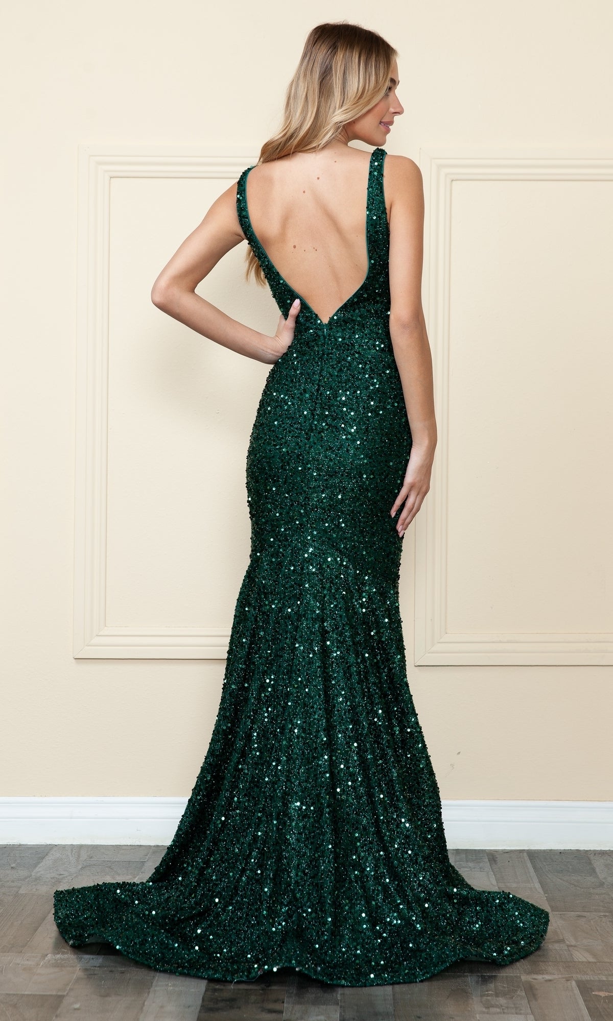A woman stands on a wooden floor facing away, wearing the V-Neck Long Sequin Prom Dress 9108—floor-length, dark green, with shimmering sequins and a deep V-back.