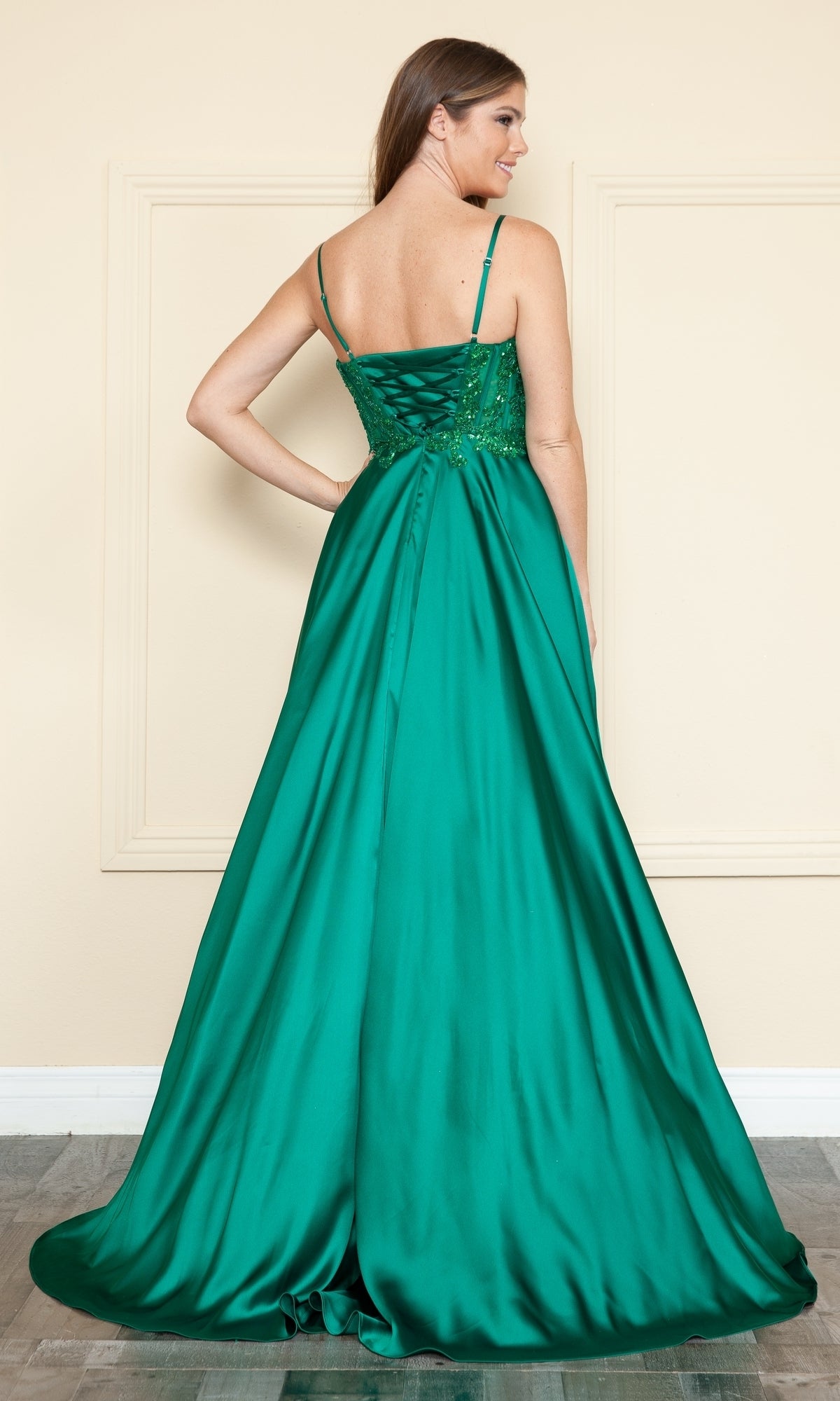 A woman stands facing away in a simple indoor setting, wearing the Sheer-Corset Long Satin Prom Dress Poly USA 9126—an emerald green, a-line gown featuring thin straps and a lace-up back.