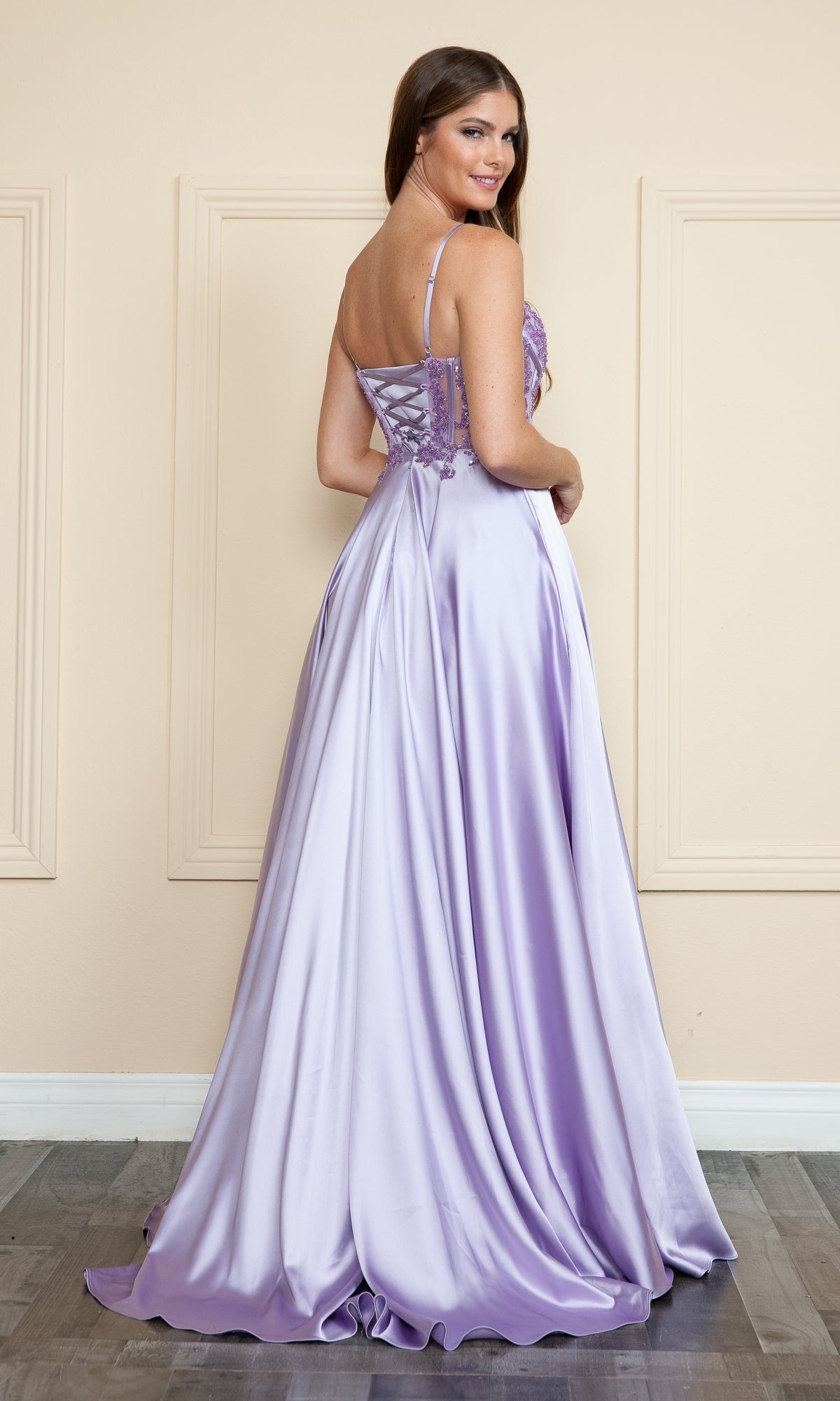 A woman stands in a room wearing the Sheer-Corset Long Satin Prom Dress by Poly USA 9126, a floor-length lavender gown with a lace-up back and embroidered corset, facing away and looking over her shoulder.