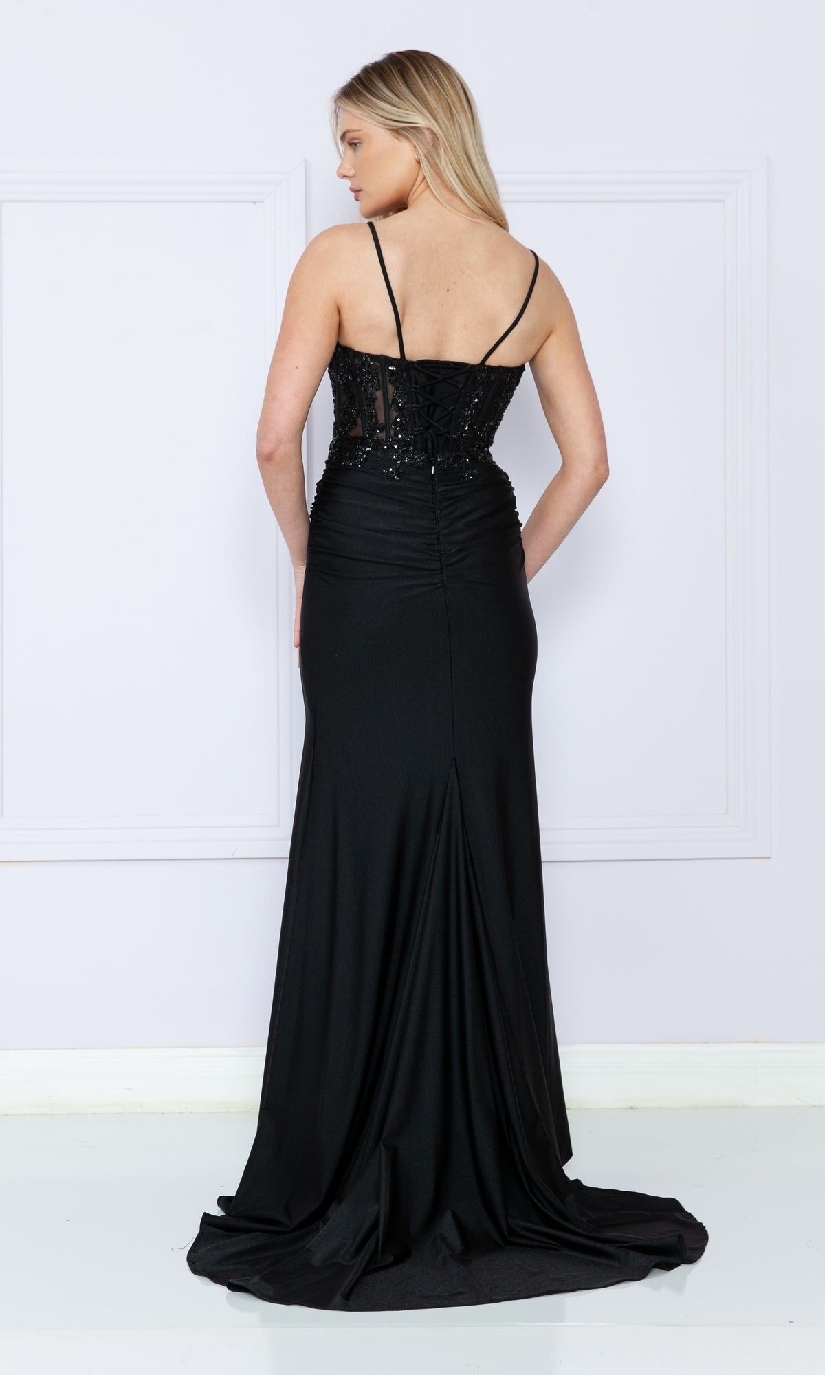 A woman stands facing away, wearing the Sheer-Bodice Long V-Neck Prom Dress 9138—a long black gown with thin straps, a fitted sheer bodice, and a graceful v-neckline.
