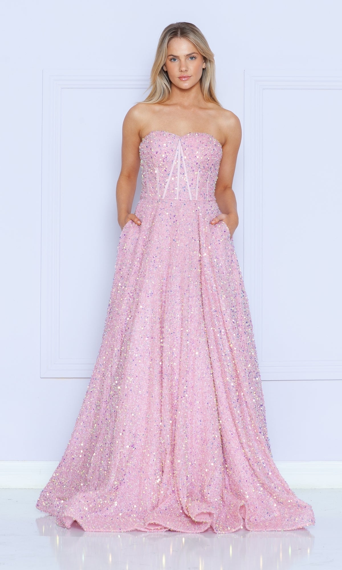 A woman stands against a pale wall wearing the Strapless Sequin Prom Ball Gown: Poly USA 9152—a floor-length, sparkly pink dress with pockets and a fitted bodice.