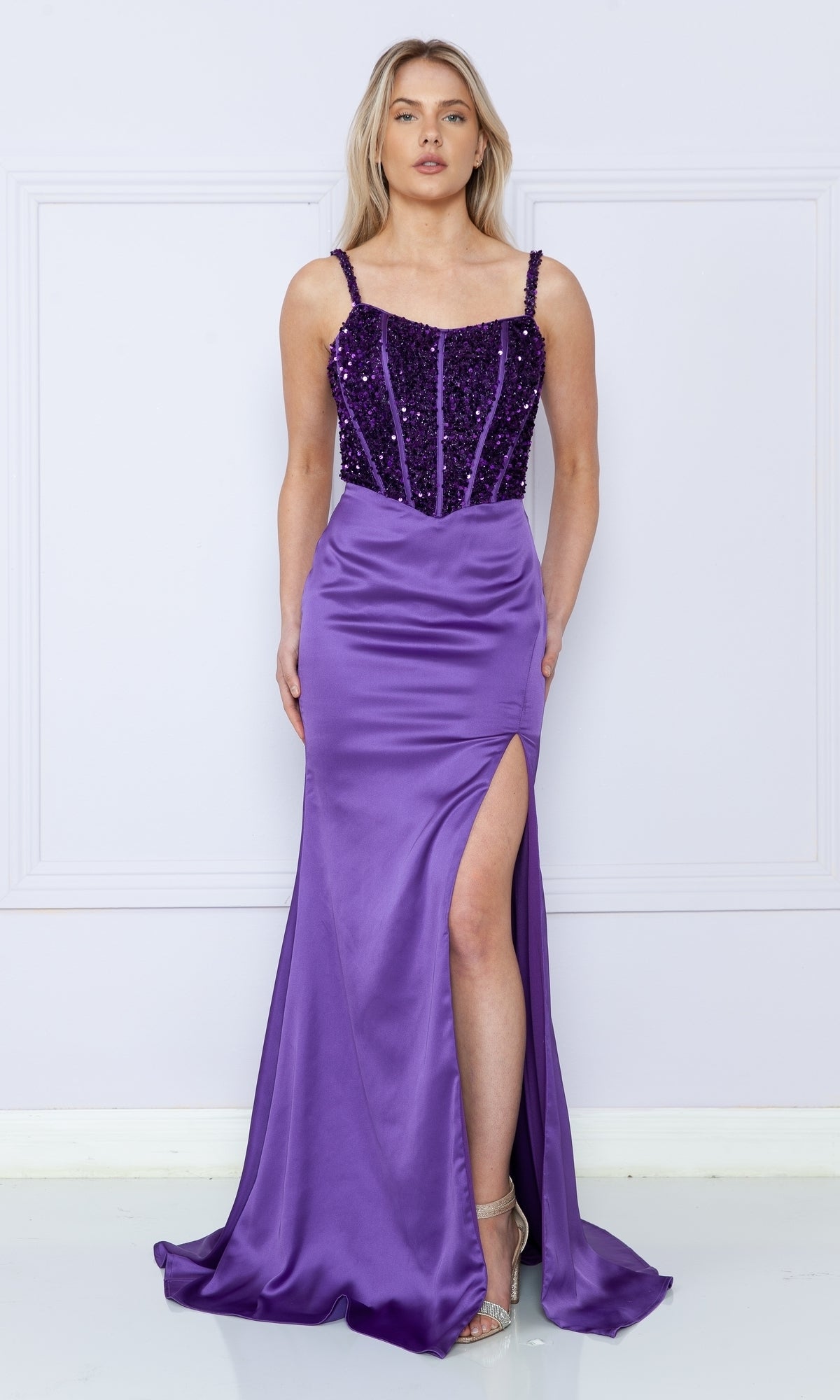 A woman in the Lace-Up Sequin-Bodice Long Satin Prom Dress 9176, featuring a sequin corset bodice, spaghetti straps, and a thigh-high slit satin skirt, stands against a light-colored wall.