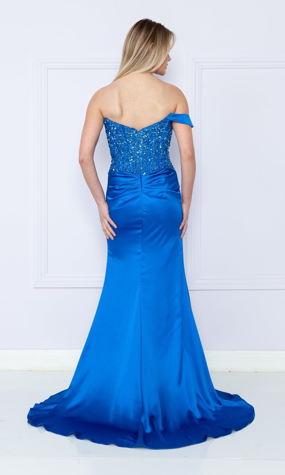 A woman stands facing away, wearing the One-Shoulder Sequin-Bodice Long Prom Dress 9178—a bright blue gown with a shimmering sequin bodice and a slight train.