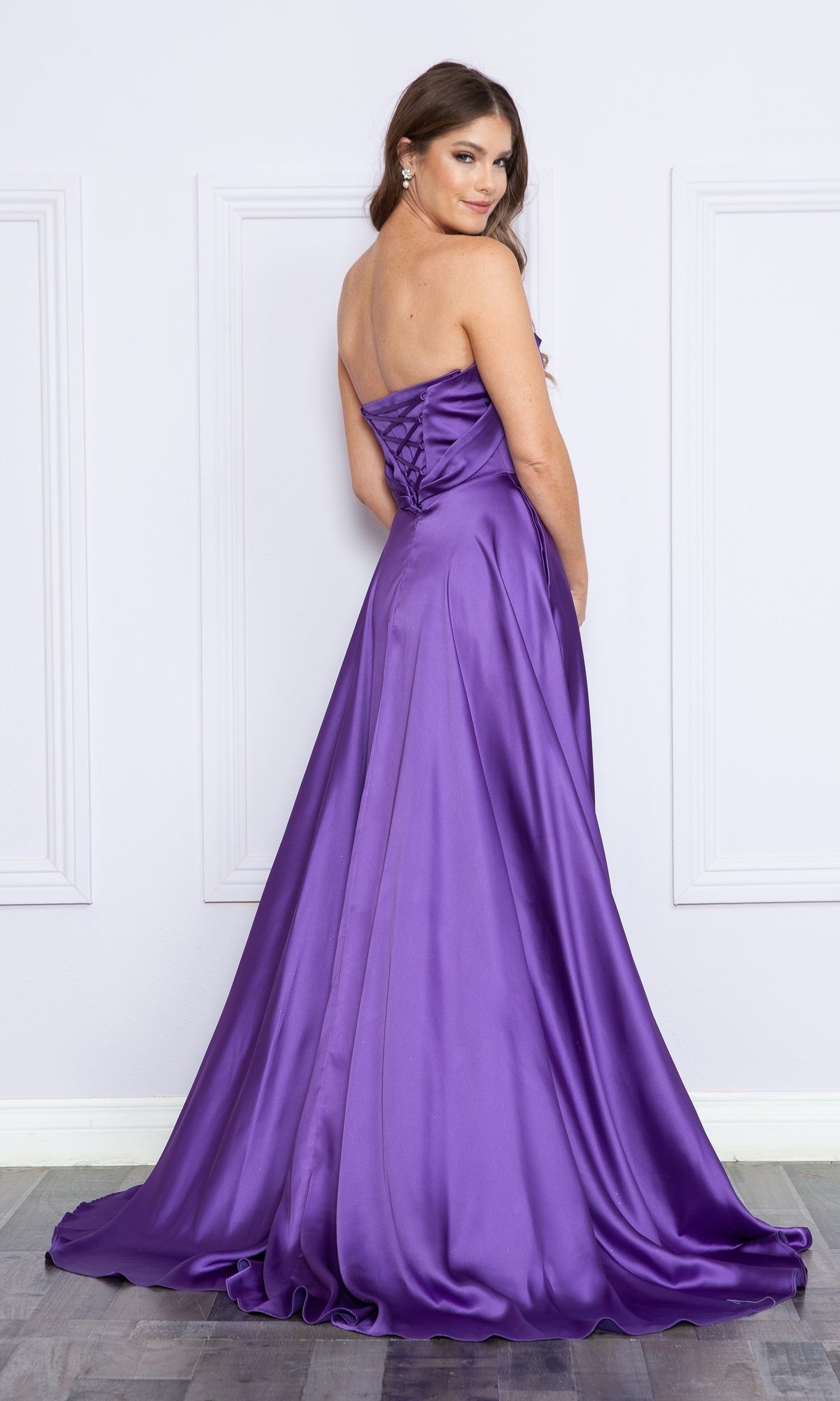 A woman, facing away and looking over her shoulder, wears a strapless long a-line prom dress with pockets; the purple satin gown features a lace-up back, floor-length hem, and a subtle side slit.