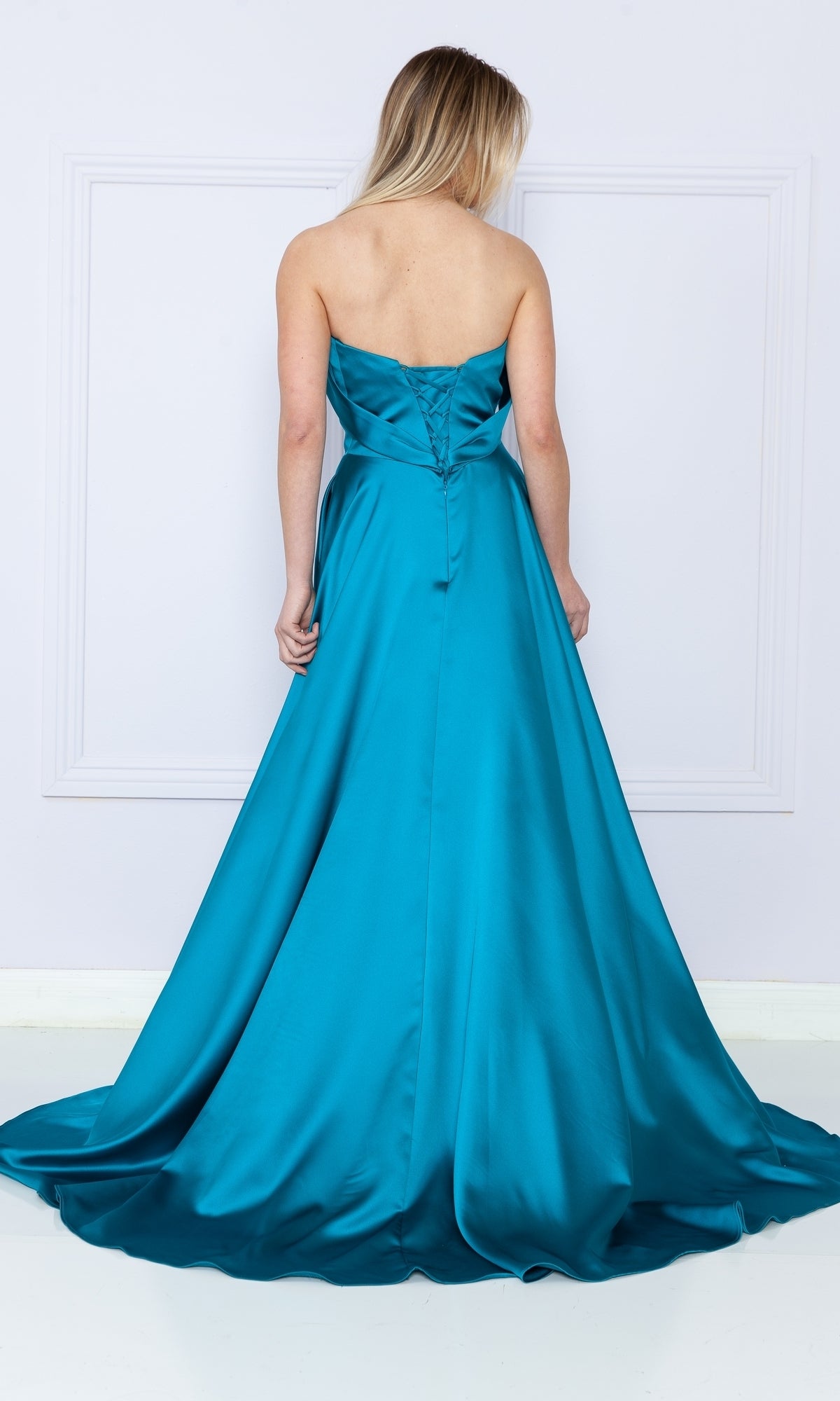 A woman stands facing away in a strapless long A-line prom dress with pockets, showing off the fitted bodice and flowing train.