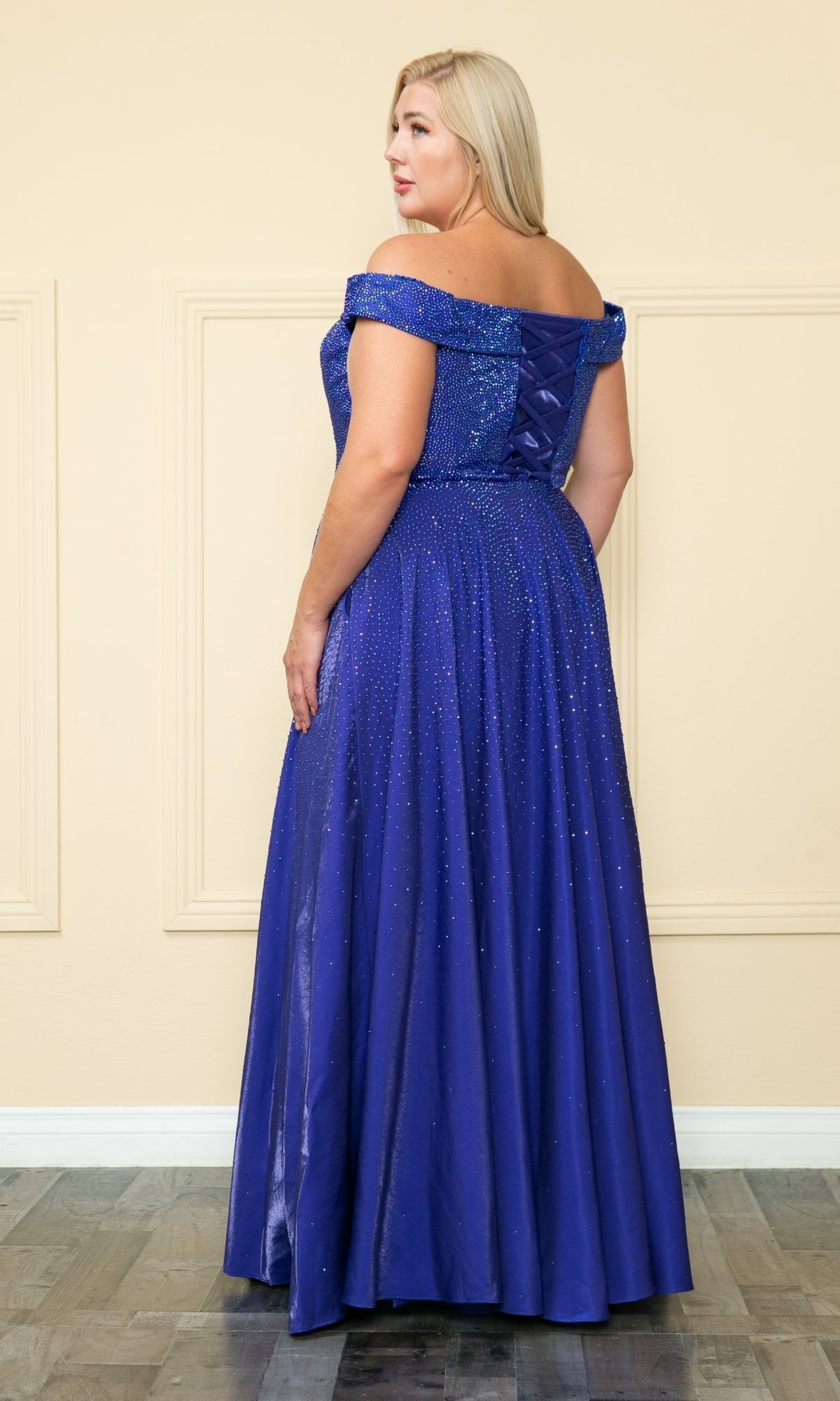 A woman stands indoors in the Plus-Size Long Beaded Prom Dress with Pockets W1112, a floor-length, off-the-shoulder gown with shimmer detail, facing slightly away from the camera.