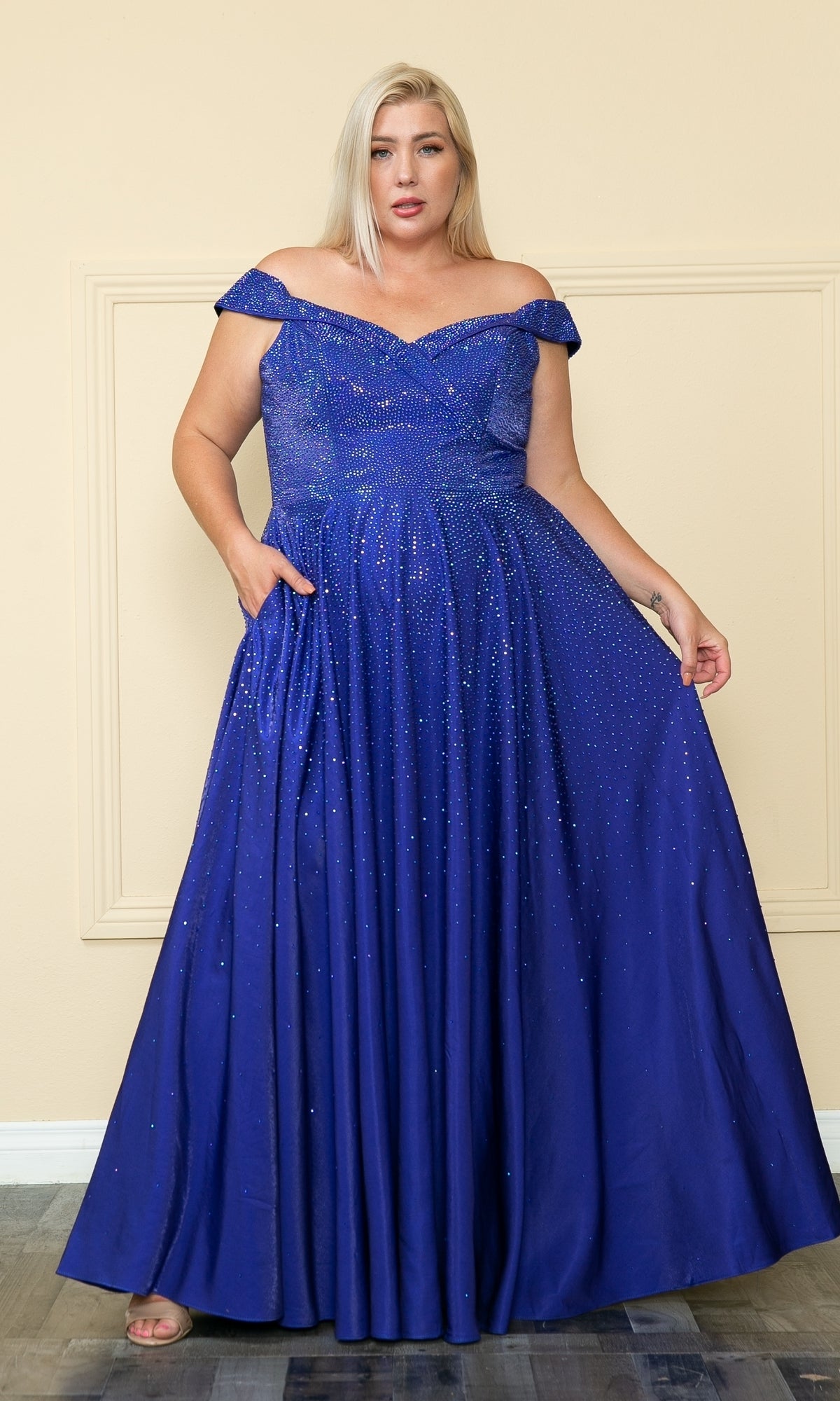 A woman stands indoors wearing the Plus-Size Long Beaded Prom Dress with Pockets W1112—a floor-length, off-the-shoulder royal blue gown with sparkling embellishments—holding out part of the skirt.