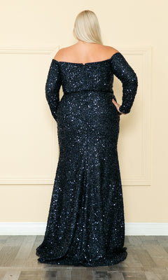 A woman with long blonde hair, seen from behind, wears the Long Sleeve Plus-Size Long Sequin Prom Dress W8876, featuring off-the-shoulder long sleeves and a floor-length silhouette, standing gracefully against a light beige wall.