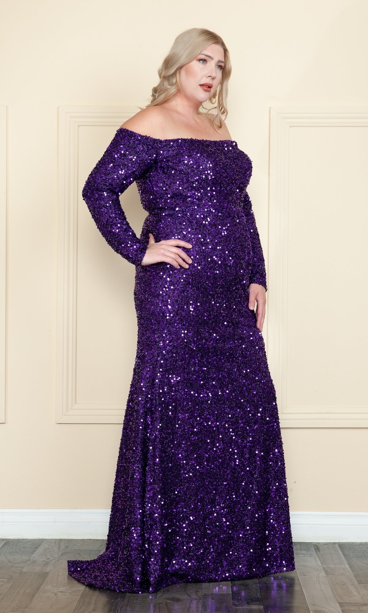 A woman stands indoors wearing the Long Sleeve Plus-Size Long Sequin Prom Dress W8876—an elegant, off-the-shoulder purple gown with long sleeves and her hand on her hip.