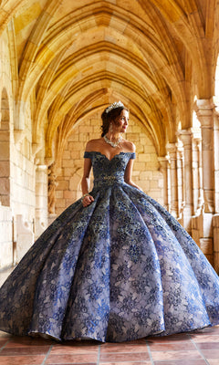 A woman in the Princesa Lace Ball Gown Quinceañera Dress PR11921 and a tiara stands in a stone archway with vaulted ceilings, gazing to the side.