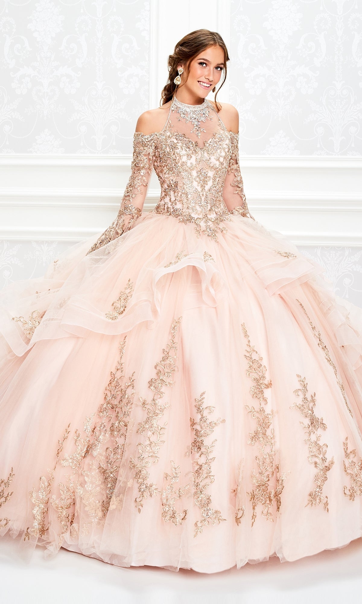 A young woman wears the Princesa Halter Ball Gown Quinceañera Dress PR11927, featuring blush pink fabric, gold embroidery, a halter neckline, and a full tulle skirt, as she stands against a white ornate background.