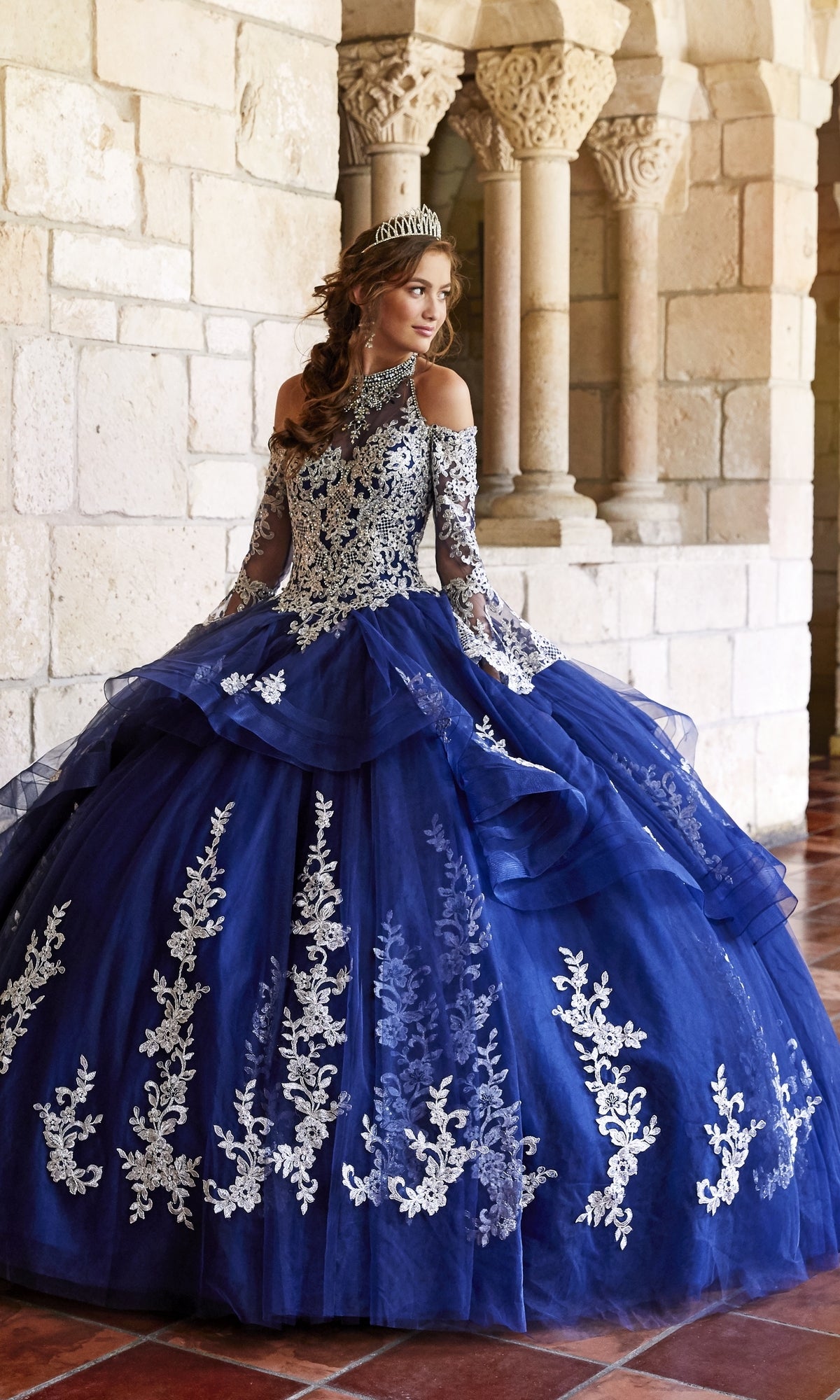 A woman wears the Princesa Halter Ball Gown Quinceañera Dress PR11927, a blue gown with white lace details and a tiara, standing in a stone hallway with columns.
