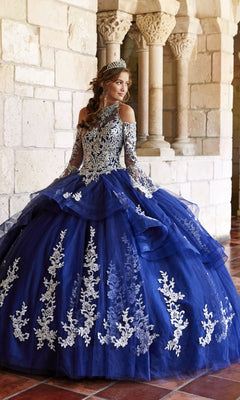 A woman wears the Princesa Halter Ball Gown Quinceañera Dress PR11927, a blue gown with white lace details and a tiara, standing in a stone hallway with columns.