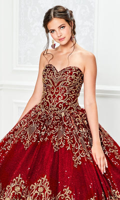 A young woman wears the Princesa Red and Gold Quinceañera Dress PR11945, a strapless ball gown with gold embroidery and sequin details, standing indoors against an ornate white background.