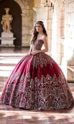 A young woman wears the Princesa Red and Gold Quinceañera Dress PR11945, standing in a stone hallway with arched doorways and checkered floor tiles.