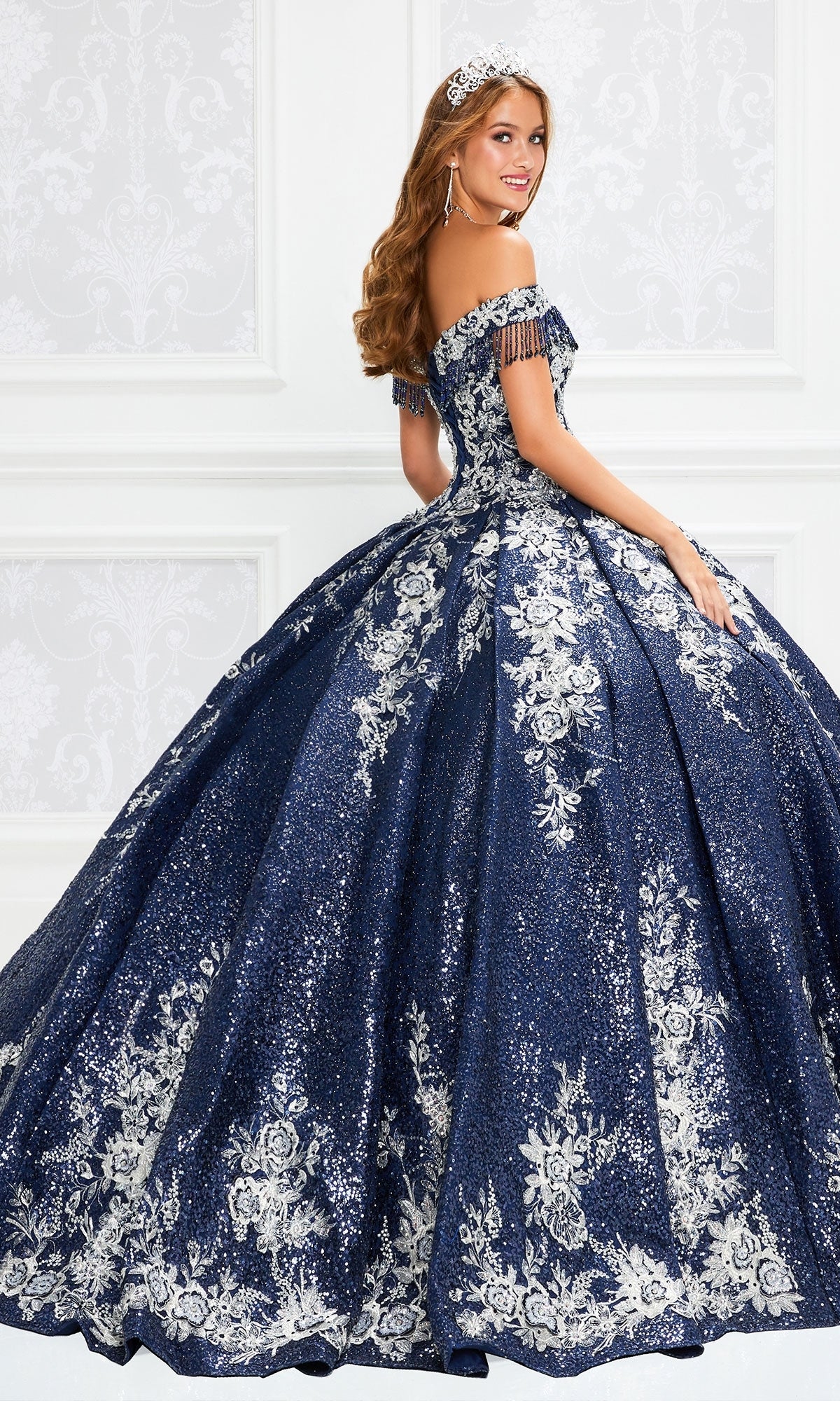 Young woman in a Princesa by Ariana Vara Quinceañera Dress PR12001, featuring off-the-shoulder design and silver floral embroidery, wearing a tiara in an ornate white room.