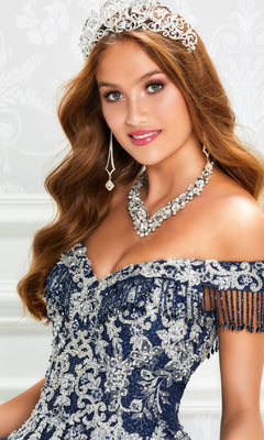 A young woman poses gracefully against a decorative white background in the Princesa by Ariana Vara Quinceañera Dress PR12001, styled with a jeweled tiara, ornate necklace, and glittering ball gown.