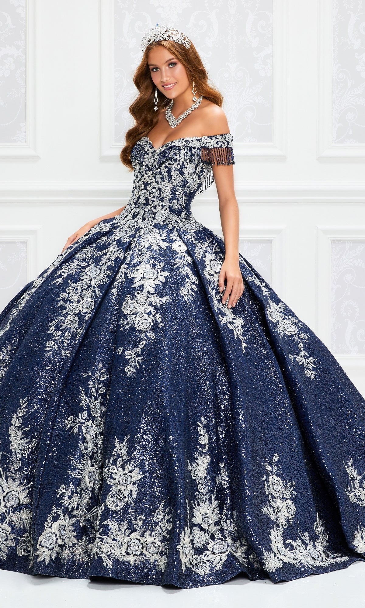 A woman stands in an elegant white room wearing the Princesa by Ariana Vara Quinceañera Dress PR12001, a glittering ball gown with silver floral details and off-the-shoulder sleeves, paired with a tiara.