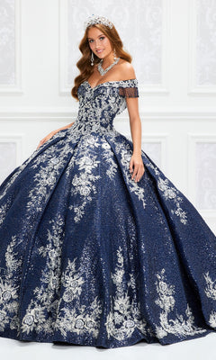 A woman stands in an elegant white room wearing the Princesa by Ariana Vara Quinceañera Dress PR12001, a glittering ball gown with silver floral details and off-the-shoulder sleeves, paired with a tiara.