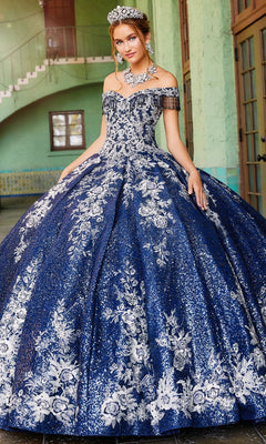 A young woman stands elegantly indoors wearing the Princesa by Ariana Vara Quinceañera Dress PR12001—a glittering off-the-shoulder gown with blue and silver floral patterns, paired with a sparkling tiara and statement jewelry.